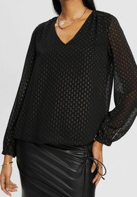 Femme portant un chemisier noir transparent à manches longues avec des pois métalliques et une jupe en cuir noire froncée, accessoirisée de bijoux en or.