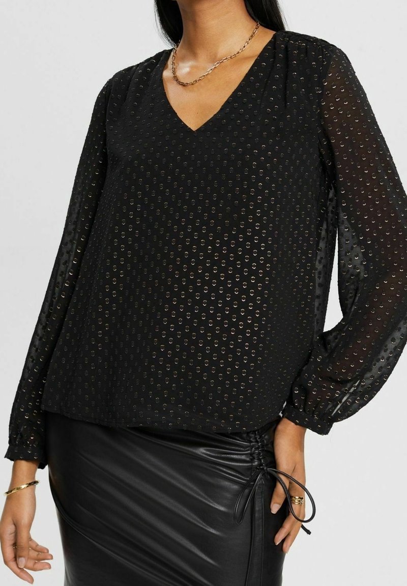 Femme portant un chemisier noir transparent à manches longues avec des pois métalliques et une jupe en cuir noire froncée, accessoirisée de bijoux en or.