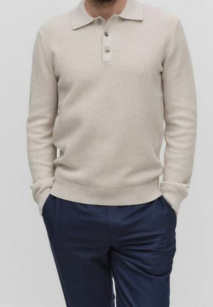 Uomo che indossa una polo beige a maniche lunghe in maglia con bottoni e pantaloni blu navy, con le mani in tasca su uno sfondo neutro.