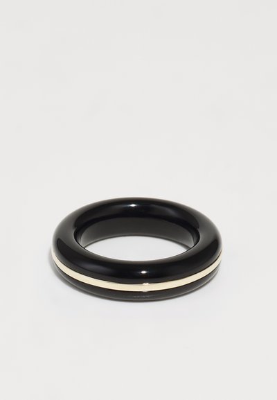 Brazalete negro brillante con una textura suave, que presenta una delgada franja de color crema alrededor de su circunferencia. Diseño circular y minimalista.