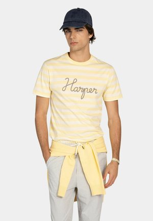 Joven con camiseta amarilla a rayas "Harper", pantalones claros, suéter amarillo anudado a la cintura, gorra azul marino y pulsera, posando frente a un fondo liso.