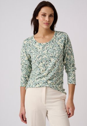 Vrouw met lang donker haar die een top met lange mouwen draagt met groen en beige bladmotief en lichte broek tegen een effen achtergrond.