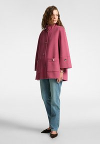 Cappotto a poncho in misto lana rosa con colletto alto, bottoni frontali e due grandi tasche anteriori. Abbinato a jeans dritti blu chiaro.