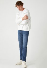 Felpa bianca, jeans blu e sneakers bianche. La felpa ha un collo rotondo e polsini a coste; i jeans sono slim-fit.