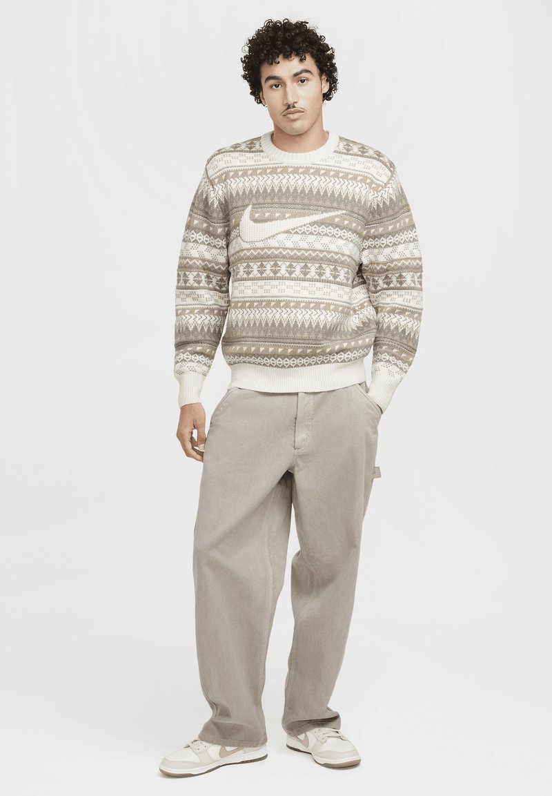 Strickpullover mit hellbraunen und cremefarbenen geometrischen Mustern, mit Rundhalsausschnitt. Kombiniert mit lockeren hellgrauen Hosen und weißen Sneakers.