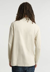 Chemise long à manches longues beige clair avec un col, présentant une texture lisse et une coupe décontractée, associée à un jean en denim foncé.