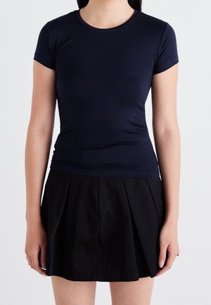 Vrouw draagt een nauwsluitend marineblauw T-shirt met korte mouwen en een zwarte geplooide rok, staand tegen een effen witte achtergrond.
