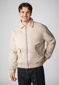 Beige Wildlederjacke mit einem Reißverschluss vorne, flachem Kragen und zwei seitlichen Taschen. Verfügt über gerippte Bündchen und Saum, getragen über dunklen Hosen.