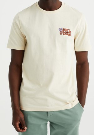 Crèmekleurig katoenen T-shirt met korte mouwen, met een bloemenprint in oranje en paars op de linkerborst.