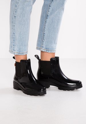 Bottes de pluie noires brillantes montantes avec panneaux latéraux élastiques portées avec un jean court bleu clair sur un sol blanc.