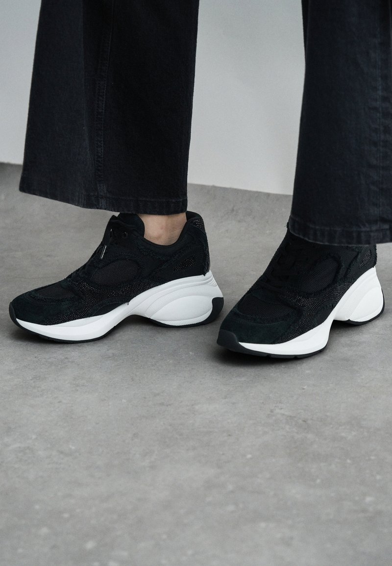 Black mesh and suede athletic shoes with a white sole, featuring a curved design and textured accents. Suitable for casual wear.