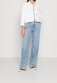 Giacca corta di jeans bianca con due tasche frontali, abbinata a jeans a gamba larga azzurri e sneakers bianche e nere.