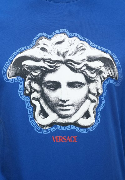 Modré bavlnené tričko s čierno-bielou grafikou Medusy, rámovanou vzorom gréckeho kľúča, s nápisom "VERSACE" červenou farbou pod grafikou.