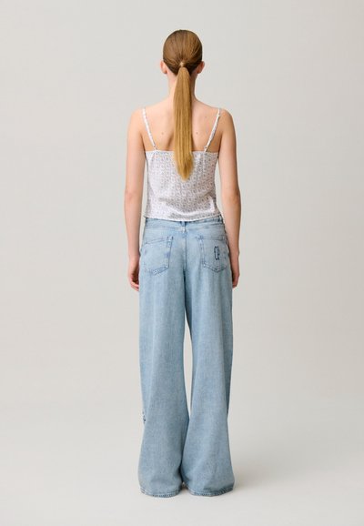 Donna con lunga coda di cavallo che indossa un top bianco floreale con spalline sottili e jeans azzurri a gamba larga, in piedi di spalle su uno sfondo semplice.
