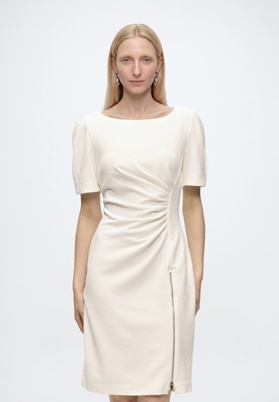 Femme aux cheveux blonds et raides portant une robe blanche texturée jusqu'aux genoux avec de courtes manches bouffantes et une fermeture éclair latérale, debout devant un fond uni.