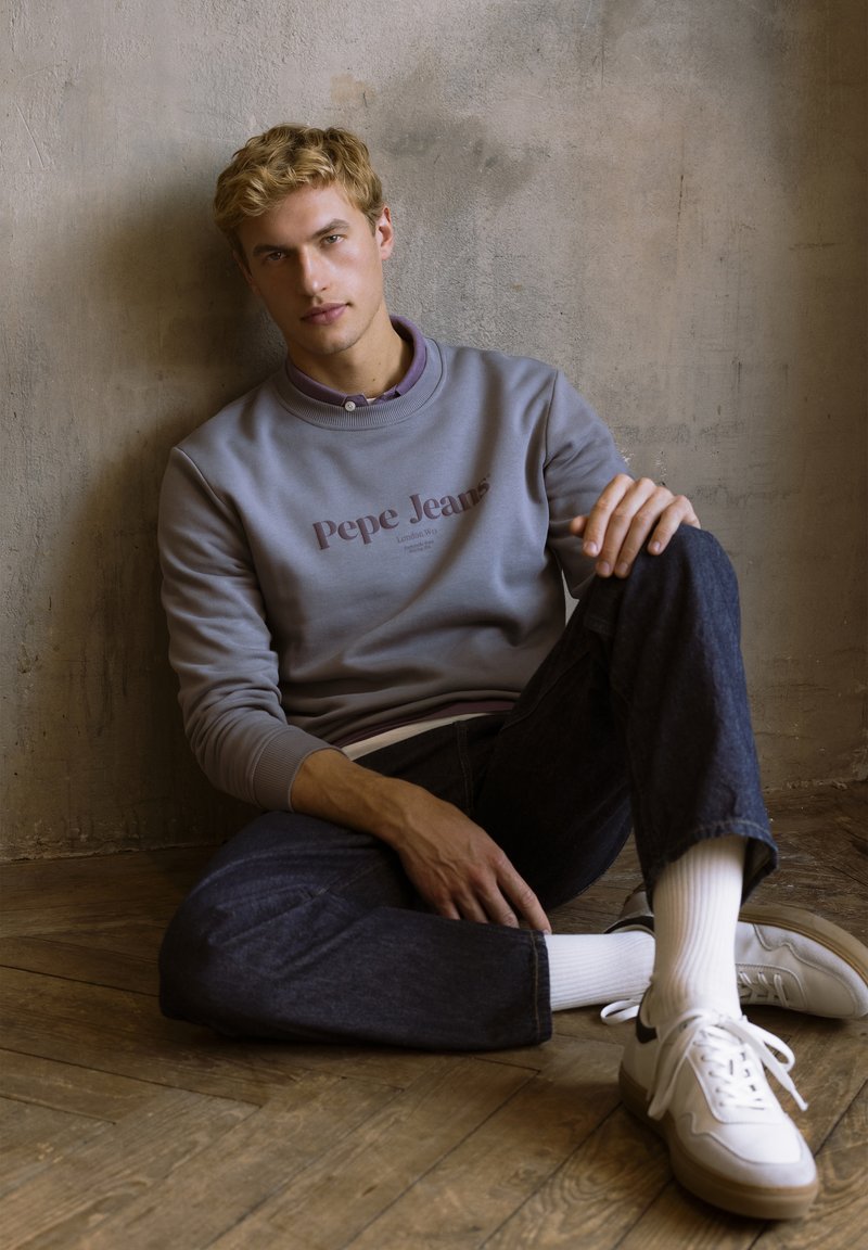 Hallhallane dressipluus, millel on "Pepe Jeans" logo, lillat värvi krae, koos tume teksapükstega ja valgete spordijalatsitega, istub puidust põrandal.