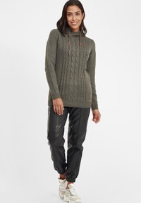 Pull-over en maille vert olive avec motif en relief, col montant avec cordons, associé à un jogging en cuir noir et des baskets beiges.