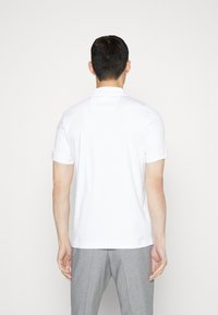 Vit pikétröja med korta ärmar, knappad krage och uppvikta ärmslut, bärs med ljusgrå byxor, sedd bakifrån.