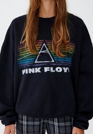Persoon die een zwart Pink Floyd sweater draagt met een prisma-ontwerp met regenboogstrepen en geruite broek, getoond van hals tot heupen.