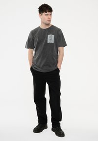 Gray cotton t-shirt with a textured black and white graphic pocket design, paired with black pants and black boots. Plain white background.
