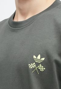 Σκούρο πράσινο βαμβακερό μπλουζάκι με κίτρινο γραφικό που απεικονίζει τρία επικαλυπτόμενα φύλλα της adidas και δύο καρό σημαίες στην αριστερή πλευρά του στήθους.