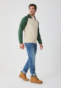 Felpa beige e verde con zip e collo a costine a contrasto, abbinata a jeans blu e stivali da lavoro color cuoio. In piedi su uno sfondo semplice.