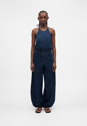 Weibliches Model mit geflochtenem Haar, trägt einen ärmellosen dunkelblauen Halter-Jumpsuit mit weitem Bein und braunen Sandalen, stehend vor einem einfarbigen Hintergrund.