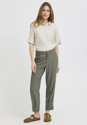 Mladá žena stojaca s rukami v vreckách, oblečená v béžovej krátkorukávovej košeli, olivovo zelených nohaviciach a hnedých slip-on topánkach so sponami.