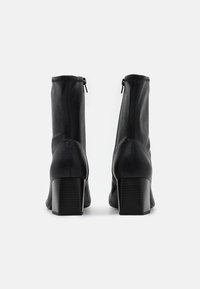 Botas de cuero negro hasta el tobillo con una textura lisa, cierre de cremallera y tacones gruesos y acanalados. El diseño presenta una silueta ajustada.