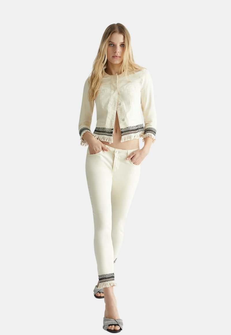 Veste en denim crème avec franges et accents à motifs noir et blanc, associée à un jean skinny crème assorti et des talons à motifs.
