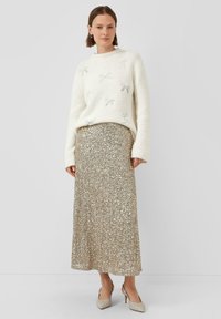 Vrouw in een witte trui met zilveren strikken, een lange zilveren paillettenrok en beige hoge hakken met slingback, staand tegen een effen achtergrond.