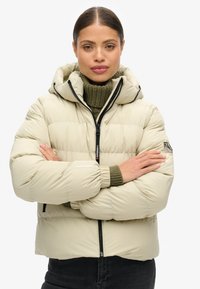 Giacca puffer beige con cappuccio, zip nera e colletto in maglia verde. Presenta un logo sulla manica sinistra e polsini elasticizzati.