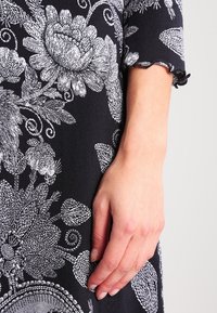 Prawa ręka oparta obok czarnego ubrania z szczegółowymi białymi wzorami kwiatowymi i falbaniastym wykończeniem rękawów.