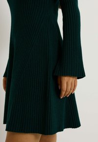 Robe à manches longues côtelées de couleur vert profond. Manches évasées et forme A. Tissu doux et texturé avec des lignes verticales et des détails de couture subtils.