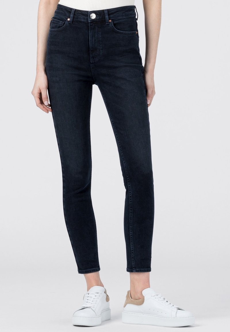 Jeans ajustados de talle alto en negro, confeccionados en denim, con un diseño clásico de cinco bolsillos y cierre de botón. Combinados con zapatillas blancas.
