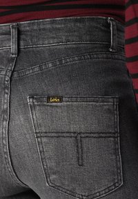 Sivé jeans z denimu zobrazujú detailný záber zadnej vrecka s logom. Tkanina má hladkú textúru a jemný lesk.