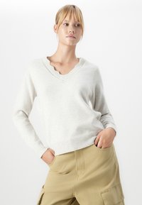 Mulher jovem com cabelo loiro, vestindo um suéter beige claro com decote em V em formato de onda e calças cargo em tom de areia, com as mãos nos bolsos.