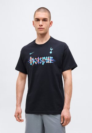 Homme aux cheveux courts portant un t-shirt noir Tottenham Hotspur avec un logo coloré et un short gris, debout devant un fond uni.