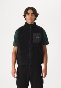 Black fleece vest with a high collar, zip closure, and a black chest pocket featuring yellow logo embroidery. Worn over a green shirt.