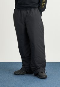 Pantaloni neri isolati con una vestibilità comoda, vita elastica, tasche laterali e cordino alla caviglia, indossati con scarpe nere su un pavimento blu.