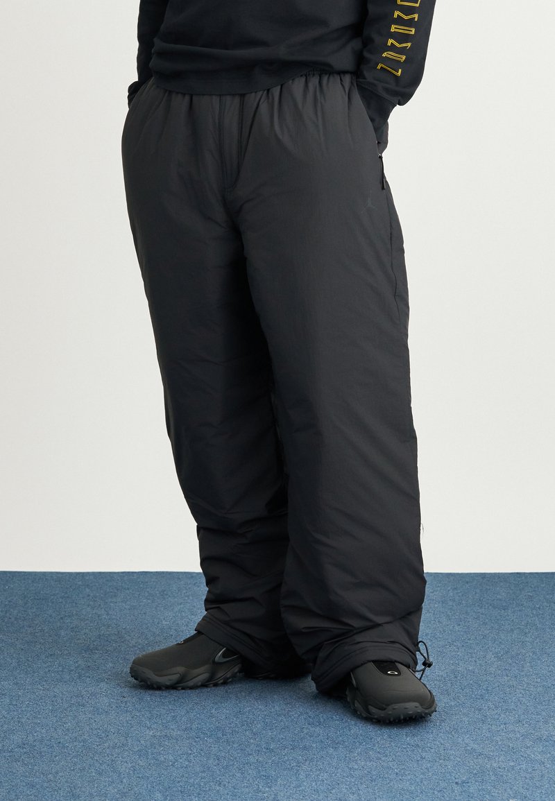 Pantaloni neri isolati con una vestibilità comoda, vita elastica, tasche laterali e cordino alla caviglia, indossati con scarpe nere su un pavimento blu.