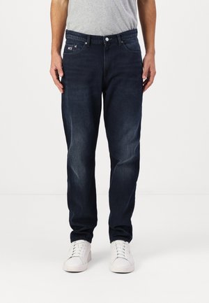 Man draagt donkerblauwe jeans met rechte pijpen en witte sneakers, staand tegen een effen witte achtergrond.