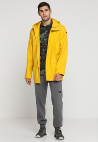 Geel waterdichte regenjas met een capuchon, drukknoopsluiting en zijzakken, gedragen over een zwart bloemenhemd en grijze joggingbroeken.