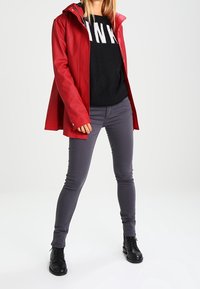 Veste imperméable rouge avec capuche, portée par-dessus un pull noir avec des lettres blanches, associée à un pantalon slim gris et des bottines noires.