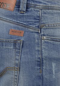 Jeans de mezclilla en un color azul desgastado, con un parche de cuero marrón etiquetado "INDICODE" y un botón y remache de metal plateado.