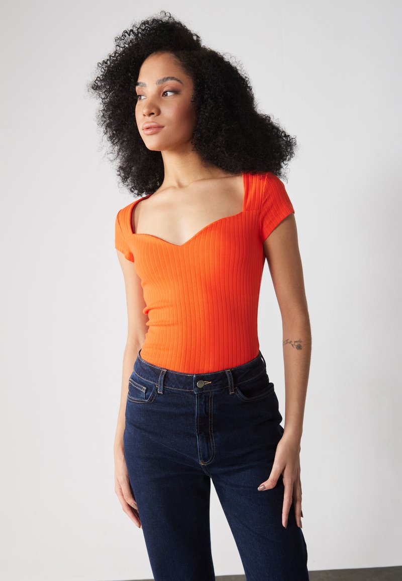 Ribbet, kortærmet bodysuit i lys orange med en sweetheart-udskæring, parret med mørke denimjeans med frontlommer og knaplukning.