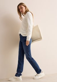 Jersey blanco y esponjoso, jeans azul oscuro y zapatillas blancas. Conjunto casual con un bolso tote de textura beige. La modelo está de pie contra un fondo claro.