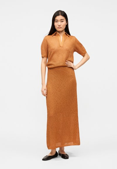 Nő, aki kezét csípőjén tartva áll, barna mintás rövid ujjú felsőt és hozzá illő hosszú szoknyát visel, fekete lapos cipővel, egyszínű háttér előtt.