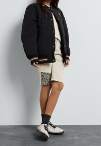 Zwarte bomberjack met gouden en witte gestreepte manchetten, crèmekleurig t-shirt, beige shorts met ruitjespatch, en grijze sokken met zwarte schoenen.