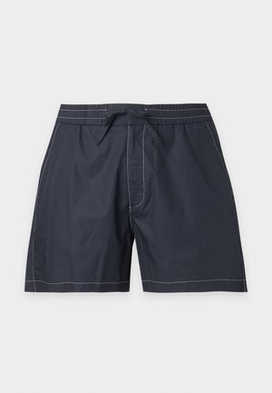 Hombre de pie con una camisa azul marino de manga corta con botones, pantalones cortos a juego, camiseta blanca y sandalias negras sin cierre sobre un fondo blanco.